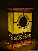 Front lit view of the stained glass mantle clock showing yellow illuminated panels, Roman numeral clock face, and decorative metal framing at base and top.