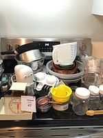 Photo showing electric hand mixer, measuring cups, metal angel food cake pans, kitchen scale, citrus juicer, egg cooker, plastic mixing bowls, and glass jars on stove top.