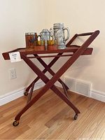 Full view of the wooden folding bar cart with wheels, holding glassware and steins on top