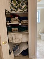 Shelving with neatly stacked assorted bedding: pillows with green leafy covers, various sheets and folded duvets. Bathroom seen in background.