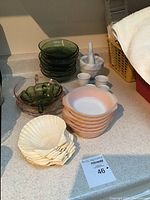 Photo of green glass bowls, shell appetizer plates, white egg cups, pink Fire King bowls, and a marble mortar and pestle on a counter.