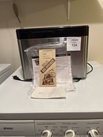 Front view of Kuraidori bread maker showing black top and stainless steel body with instruction manual and accessories on counter in front.