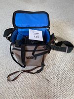 Open beige padded camera carry bag with blue interior and black adjustable shoulder strap, sitting on carpeted surface. A paper tag with lot number is placed inside the bag.