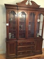 Front view of the solid wood china cabinet showing four glass doors, wooden base with three drawers and two cupboards, ornate trim and molding on top.