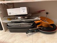 Overview of assorted cookware stacked on floor including pans, trays, a yellow plastic handled item, and a metal wire basket shaped like a fish.