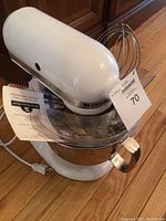 White KitchenAid Artisan mixer assembled with stainless bowl and wire whip on hardwood floor