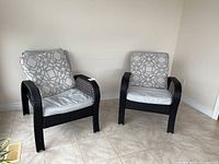 Two black resin outdoor chairs with light gray and patterned fabric cushions, placed on tiled floor near white walls.