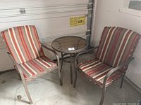 Photo showing two metal framed outdoor chairs with red and beige striped cushions alongside a round glass top patio table with matching metal frame.