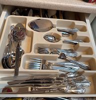 Photo of a plastic cutlery tray containing various silver-tone metal forks, knives, spoons, and a metal vegetable cutter/spatula inside a kitchen drawer.