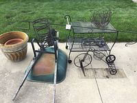 Photo of the green metal wheelbarrow with wooden handles next to the black metal garden cart with two wire baskets and two large wheels on concrete with grass in background.