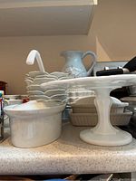 Overview of white cookware and dishes including pitcher, ladle, covered dish, stack of bowls, and cake plate on countertop.