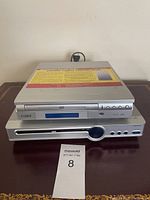 Two silver DVD players stacked on a table, showing front view of Coby and Desay players, power cord visible behind.