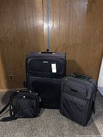 Set of three black luggage pieces: large upright suitcase, medium upright suitcase, and a small shoulder bag, on carpet with a wood-paneled wall background