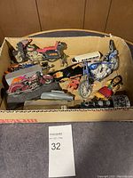 Top view of assorted toy vehicles and figurines in a cardboard box showing motorcycles, limousine, train engine, and figures.