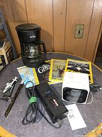 Wide view of lot showing Mr. Coffee drip coffee maker, National Geographic magazines, black power adapter with cord, sharpening stone and small tool on table against wood panel wall.