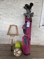 Golf club bag with clubs, wooden tennis racket leaning against wall, two soccer balls on the floor