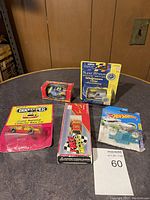 Five new, sealed diecast and collectible cars on table: Super Wheels Walgreens 100th Anniversary truck, Hot Wheels car, Innkeeper Grand National Electric Race Car, red car in clear box, white car in Ride Makers packaging.