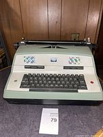 Front view of IBM electric typewriter with keyboard, IBM logo, and holographic decals visible.
