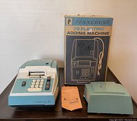 The Penncrest 78 electric adding machine in blue with white keypad next to the original box and two accessories: an orange cloth cover and a blue plastic cover.