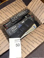 Box containing eight various remote controllers of different sizes, colors, and button layouts. Some have brand logos like JVC and GE.