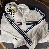 Photo showing a folded white Adidas soccer jacket with blue stripes and a Samsung logo, along with a white Real Madrid cap embroidered with the team emblem and crown.