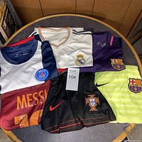Top view of five soccer jerseys including PSG, Real Madrid, and multiple FC Barcelona jerseys, and a pair of black Portugal shorts with Nike logo on a round table.