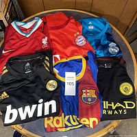 Photo showing six different soccer jerseys from well-known football clubs in various colors and sizes laid out on a round table.