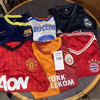 Six soccer jerseys laid out on a round table visible showing club crests and player name on one jersey.