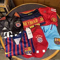 Photo showing five soccer jerseys from Bayern Munich, Barcelona, and Manchester United in varying colors and styles.