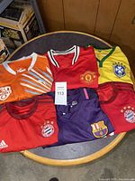 Photo showing a collection of six of the soccer jerseys arranged on a round table. Jerseys include Manchester United (red), Brazil national team (yellow with green trim), Bayern Munich (red), Barcelona (purple and red), and an orange Puma jersey with white and light blue stripes.
