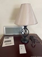 Table lamp with decorative twisted metal base and beige lampshade, power cord coiled beside it, and a Timex digital clock behind with user guide in front.