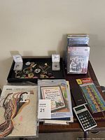 Full lot overview showing VHS tapes stacked vertically near the wall, a black tray with assorted jewelry including bracelets, necklaces, and earrings, and stacks of cross stitch kits and patterns with colorful floss.