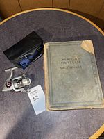 Photo showing a worn Webster's Universal Dictionary and a fishing reel with carrying case on a round wooden table.