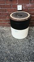 Front view of the 6 gallon stoneware crock with lid against a brick background.