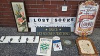 All decorative items displayed on ground against brick wall, including framed floral artwork, humorous and inspirational signs, coat rack, decorative dish, and small prints.