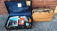 Open suitcase containing multiple JFK related magazines and booklets, next to a tan bag holding more items.