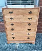 Front view of a blonde wood chest of drawers with six drawers and dark metal handles, placed outdoors on pavement against a window and brick wall.