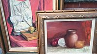 Close view of the two framed oil paintings leaning against an exterior brick wall. One depicts an abstract impressionist sculpture of a horse figure on a book with red drapery, the other a still life with vase and vegetables.