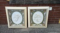 Two framed relief artworks showing angels and cherubs in oval shapes with green and gold ornamental borders, resting on pavement outdoors.