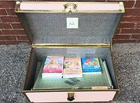 Storage trunk open showing four Barbie VHS/DVD tapes inside on top of green mat.