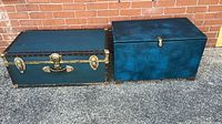 Two vintage storage trunks side by side on pavement with brick wall background. Deep blue trunk with brass hardware on left, mottled teal trunk with simple latch on right.