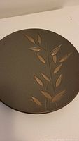 Top view showing dark gray/brown round plate with embossed tan leaf design.