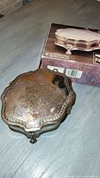 Pure silver jewelry box with a scalloped top and ornate feet resting on a grey surface. The lid shows slight tarnish. Shown alongside the original box.