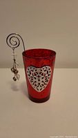 Photo showing the red candle holder with silver heart design and attached metal stick with hanging charms next to it.