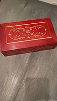 Red wooden musical jewelry box top showing detailed gold decorative design.