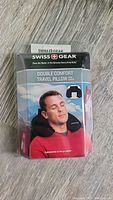 Front view of the Swiss Gear Double Comfort Travel Pillow sealed in original packaging, showing a man wearing the neck pillow on the front.