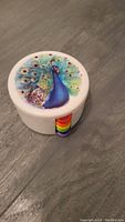 Single round plastic coaster with colorful peacock illustration and rainbow colored handle, placed on gray surface.