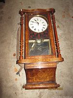 Full view of antique wooden wall clock showing intricate inlaid marquetry and turned side columns, clock face at top, and base with mirror panel below pendulum compartment.