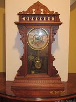 Front view of the wooden mantel clock showing ornate carved top, scrollwork on sides, Roman numeral dial and pendulum through glass door.