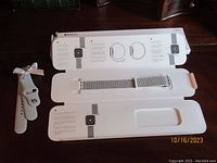 White silicone sport band and silver Milanese loop Apple Watch bands laid inside opened packaging, showing products and packaging details.